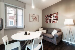 A modern living room with a round white table, two white chairs, a grey sofa with white cushions, a framed rooster artwork, an abstract painting, and a floor lamp.