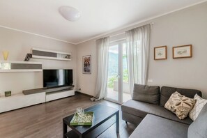 A modern living room with a flat-screen TV, a sofa, a coffee table, and a balcony with curtains. - Shimmering Waters - My Home In Como (MOLTRASIO)