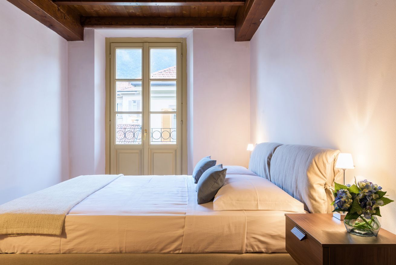 A hotel room with a bed, a nightstand with a lamp, a window with a view of buildings, and a wooden ceiling.