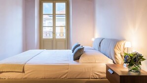 Habitación de hotel con cama, mesita de noche con lámpara, ventana con vista a edificios y techo de madera.