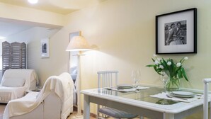 A dining area with a glass table, white chairs, a lamp, and a framed photo on the wall.