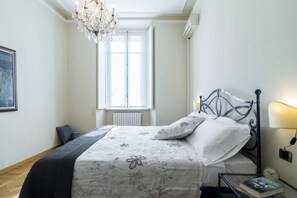 A bedroom with a bed, a chandelier, a window, a radiator, a bedside table with a lamp, and a framed picture on the wall. - Palazzo Garibaldi - My Home In Como (Como)