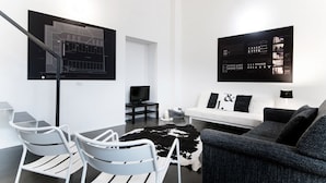 Ein modernes Wohnzimmer mit einem weißen Sofa, einem schwarz-weißen Kuhfellteppich, einem grauen Sofa, einem Esstisch mit Stühlen und zwei schwarzen Bilderrahmen mit Architekturzeichnungen an der Wand.