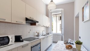 Ett modernt kök med vita skåp, en mikrovågsugn, en spis och en diskho. Det finns ett matbord med en frukostuppsättning och ett fönster med utsikt över omgivningen.