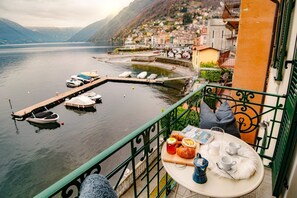 A balcony with a table set for breakfast overlooking a lake with boats and a hillside town.