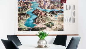 Una mappa incorniciata del Lago di Como su una parete, un tavolo rotondo con una pianta in vaso e quattro sedie nere.