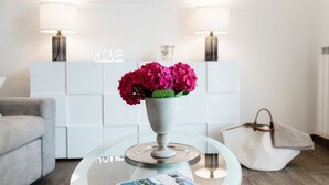 Un salon moderne avec une table basse en verre, un livre, un vase de fleurs, un canapé, un meuble blanc orné de l’inscription « HOME » et un sac à main.
