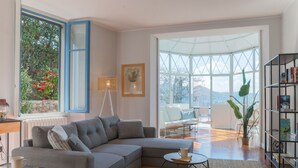Ein modernes Wohnzimmer mit einem grauen Ecksofa, einem kleinen runden Couchtisch, einem Bücherregal und einem großen Fenster mit Blick auf Bäume und Berge.