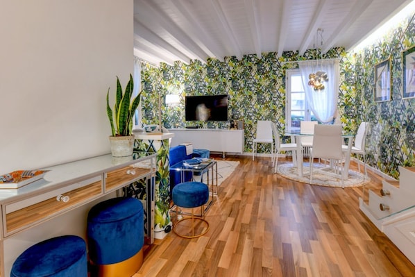 A modern living room with a wooden floor, a TV, a dining area with chairs, and a patterned wall.