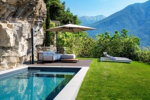 A swimming pool with a stone wall, a lounge area with a table and chairs, and a sunbed.