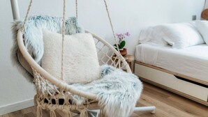 Ein Zimmer mit einem weißen Bett, einem Hängestuhl aus Makramee mit einem flauschigen Kissen und einem kleinen Topfpflanz auf einem Nachttisch.
