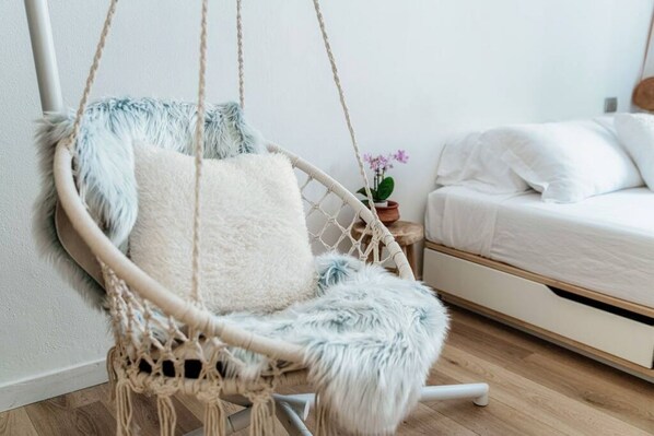 A room with a white bed, a macramé hanging chair with a fluffy cushion, and a small potted plant on a nightstand.