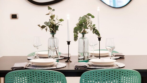 A dining table set with plates, glasses, and candles, flanked by two vases with greenery.
