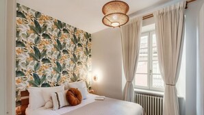 A bedroom with a floral wallpaper, a bed with white bedding, a bedside lamp, and a window with curtains.