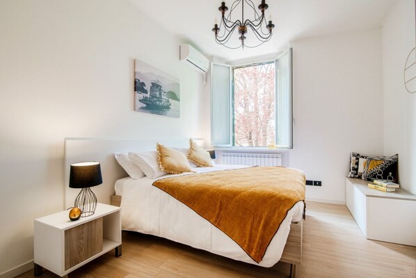 A bedroom with a bed, bedside table, lamp, window, and artwork.