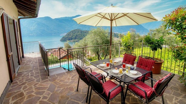 A terrace with a table set for dining, a large umbrella, and a view of a lake and mountains.