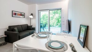 Un espace repas moderne avec une table blanche dressée pour deux personnes, un canapé foncé et une grande fenêtre donnant sur un paysage verdoyant.