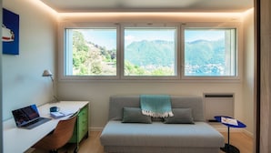 Ein Schlafzimmer mit Bett, Schreibtisch mit Laptop, Stuhl und Blick auf Berge durch große Fenster.