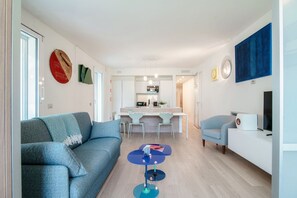 Una sala moderna con un sofá, una mesita, un comedor con sillas, una cocina al fondo y un televisor sobre un mueble blanco.