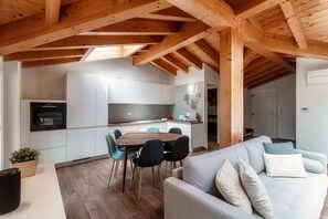 Una cocina moderna con armarios blancos, un comedor con mesa y sillas de madera, y una sala de estar con sofá y cojines.