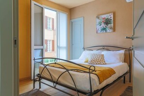 Quarto com estrutura de cama em metal, roupa de cama branca, manta amarela e porta que dá para área externa.