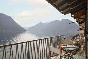A balcony with a view of a calm lake and mountains, a chair, and a table with a book and a blanket. - La Casa del Pescatore - MyHomeInComo (Colonno)
