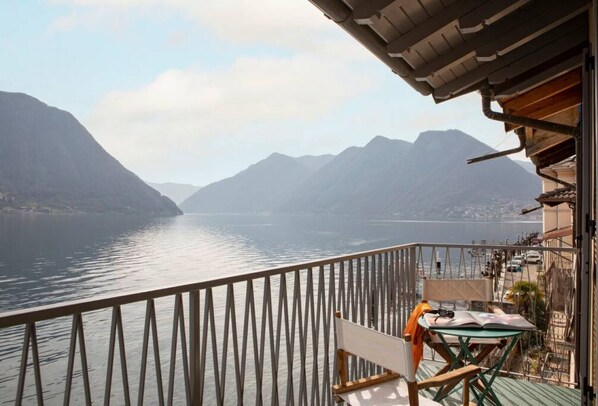 A balcony with a view of a calm lake and mountains, a table with a chair, and a building with a sloped roof. - La Casa del Pescatore - MyHomeInComo (Colonno)