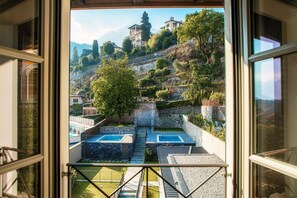 Blick vom Balkon auf einen Pool, eine Steinmauer und eine Hügelkuppe mit Gebäuden.