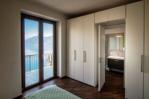 Un dormitorio con un amplio ventanal corredera que da a un balcón con vistas a montañas y un lago.