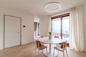Quarto moderno com mesa de jantar redonda, quatro cadeiras, luminária no teto, porta e uma grande janela com cortinas.