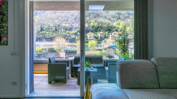 Ein Zimmer mit einer Schiebeglasdoor zum Außenbereich mit Patio, Korbmöbeln und Blick auf eine Hügelkulisse mit Gebäuden.
