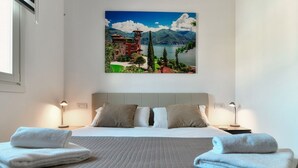 Ein Hotelzimmer mit einem Bett, zwei Nachttischen, einem Bild von einem See und Bergen sowie einem Fenster.