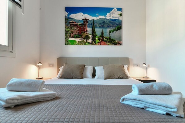 Una habitación de hotel con una cama, dos mesitas de noche, un cuadro de un lago y montañas, y una ventana.