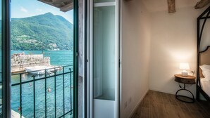A balcony with a view of a lake and mountains, a wooden floor, a bed with a headboard, and a nightstand with a lamp. - Acqua - My Home in Como (Moltrasio)