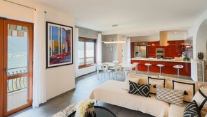 A modern living room with a sofa, a dining area with chairs, a kitchen with red cabinets, and a balcony with a view.
