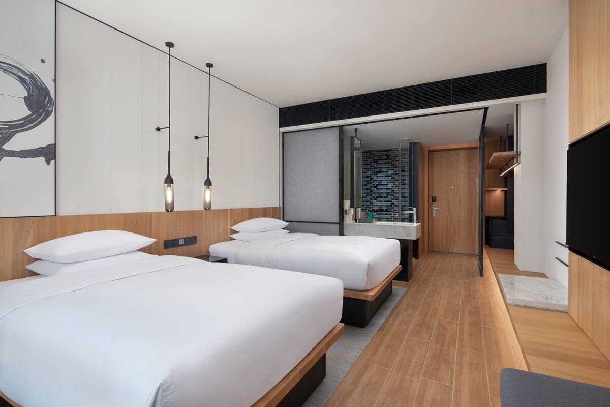 Une chambre d’hôtel moderne équipée de deux lits, d’un grand miroir, d’une télévision et d’une salle de bain avec douche et lavabo.