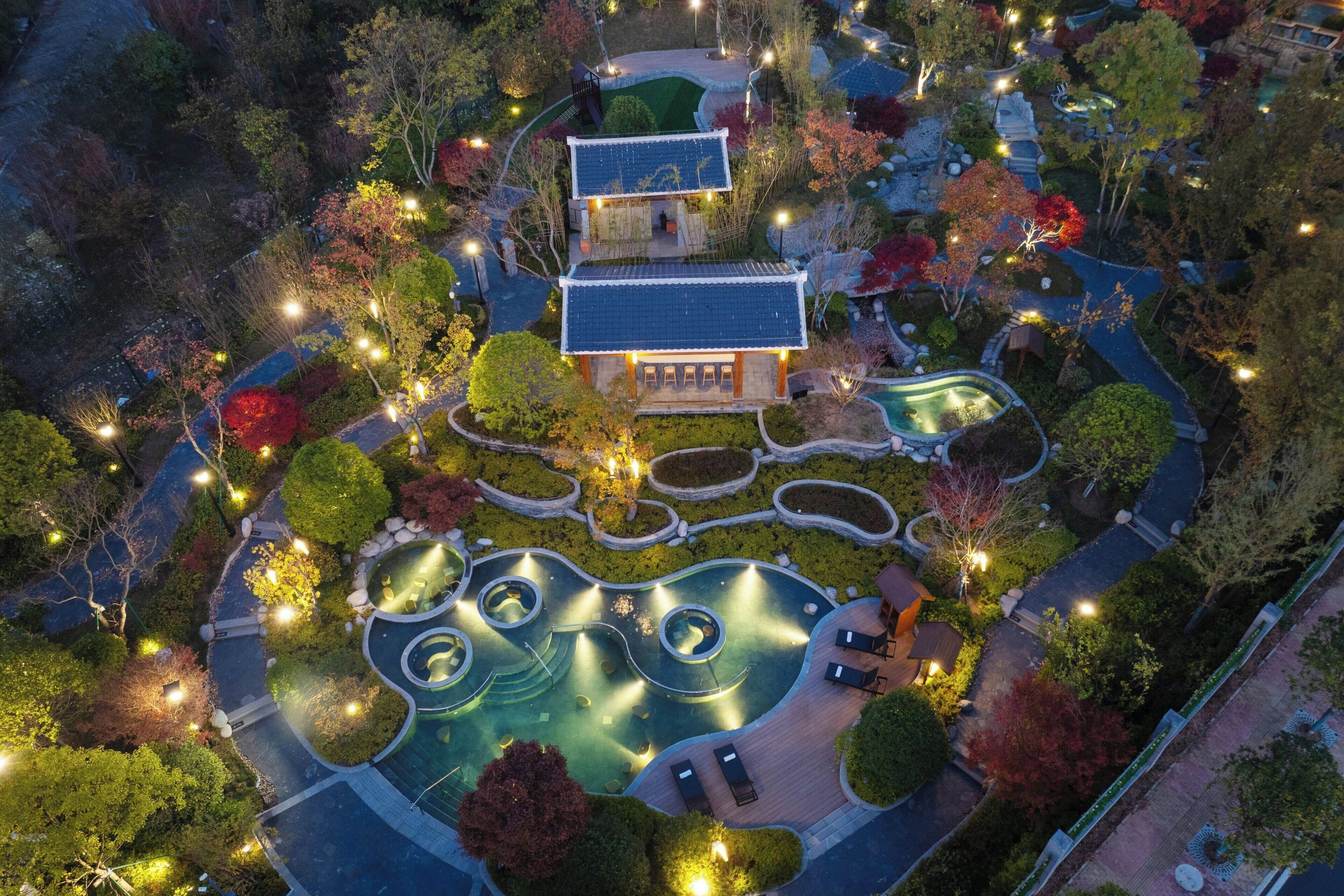 An aerial view of a traditional Japanese-style building surrounded by landscaped gardens, including a circular pool area and a small bridge.