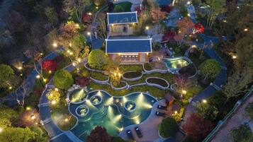 Vue aérienne d’un bâtiment de style japonais traditionnel, entouré de jardins aménagés, comprenant un espace avec une piscine circulaire et un petit pont.