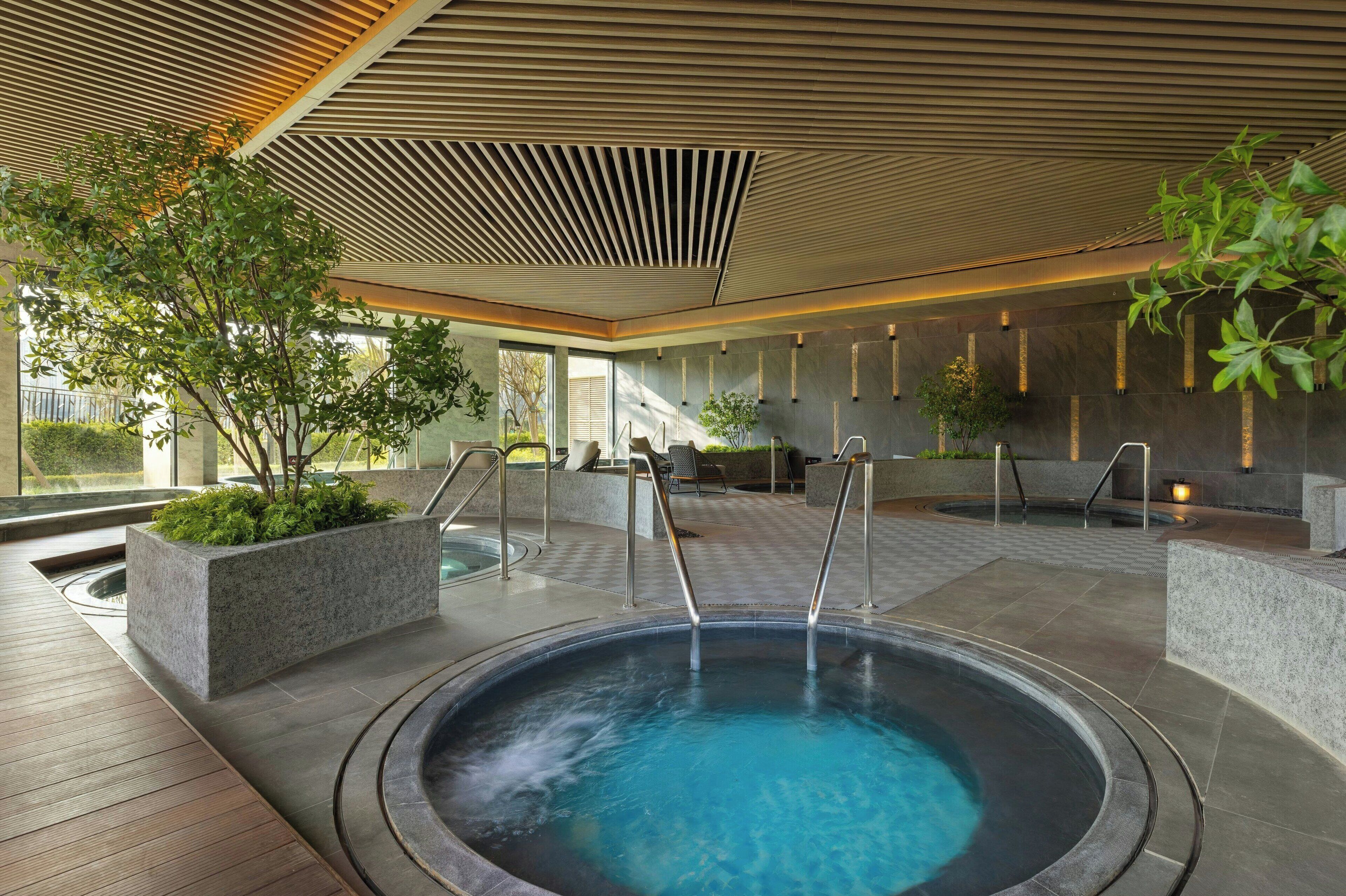 A modern indoor pool area with a hot tub, seating areas, and planters with greenery.