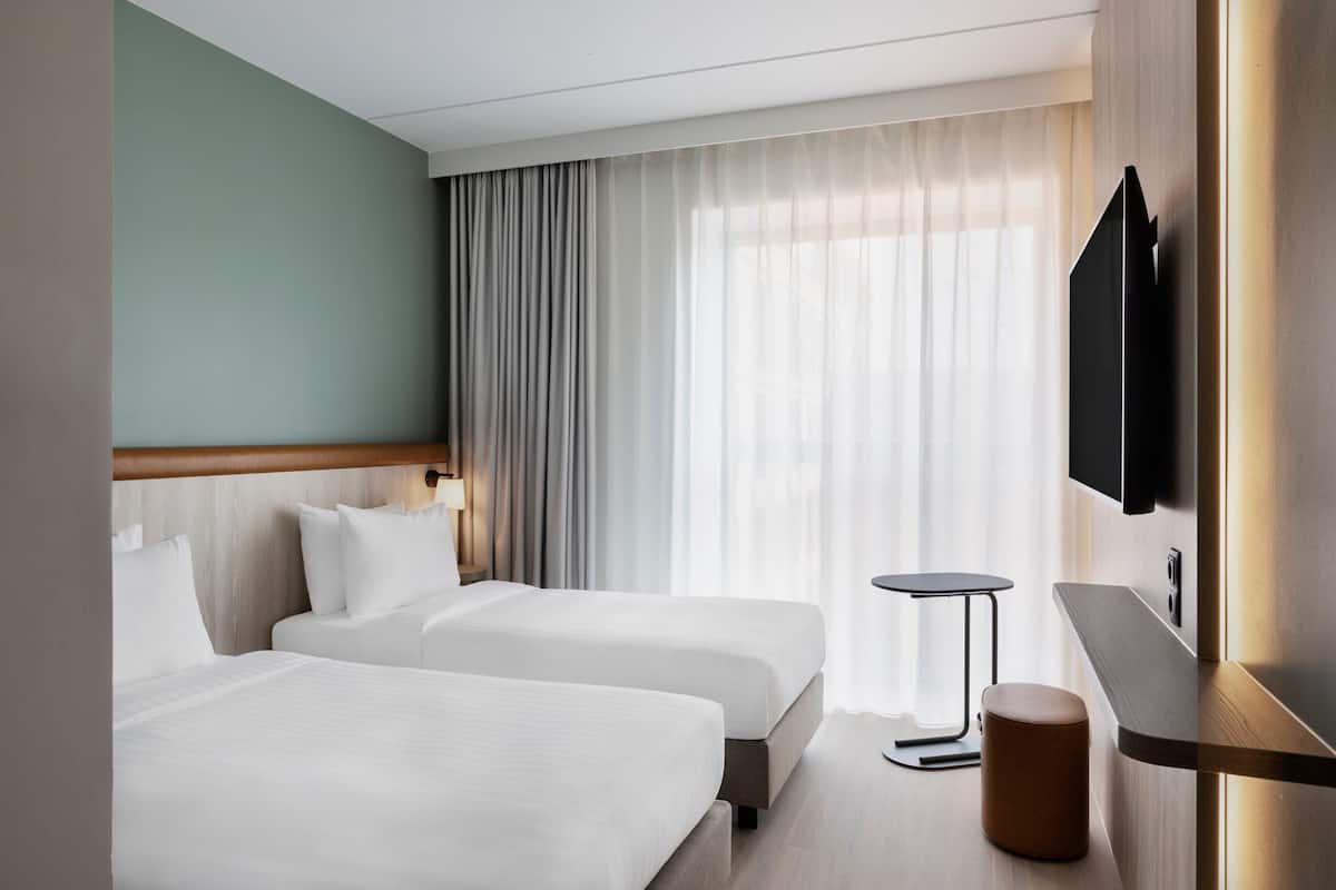 Une chambre d’hôtel avec deux lits, une petite table ronde, un téléviseur fixé au mur et une grande fenêtre avec des rideaux.