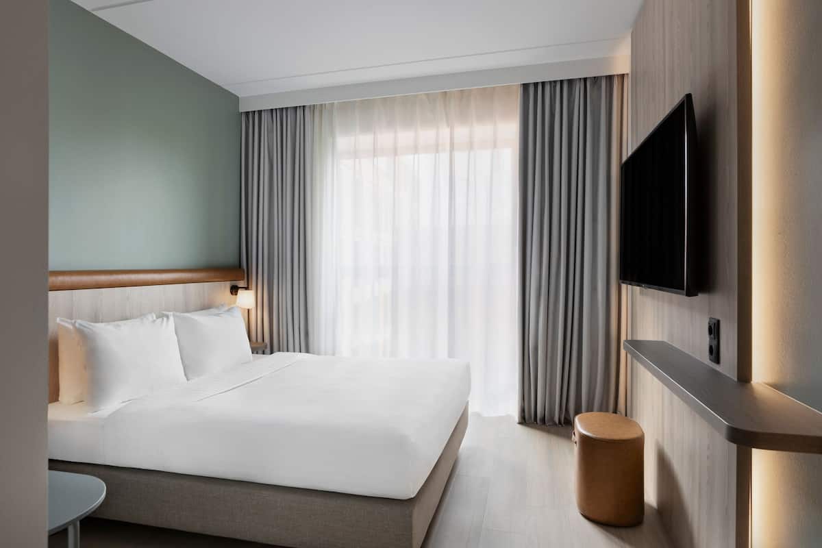 Une chambre d’hôtel moderne avec un grand lit, une petite table ronde, un téléviseur à écran plat fixé au mur et une grande fenêtre avec des rideaux.