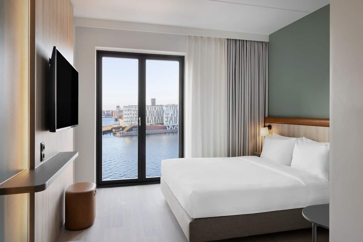 Une chambre d’hôtel avec un grand lit, un téléviseur fixé au mur, une vue sur le paysage urbain à travers une porte vitrée et une petite table ronde.