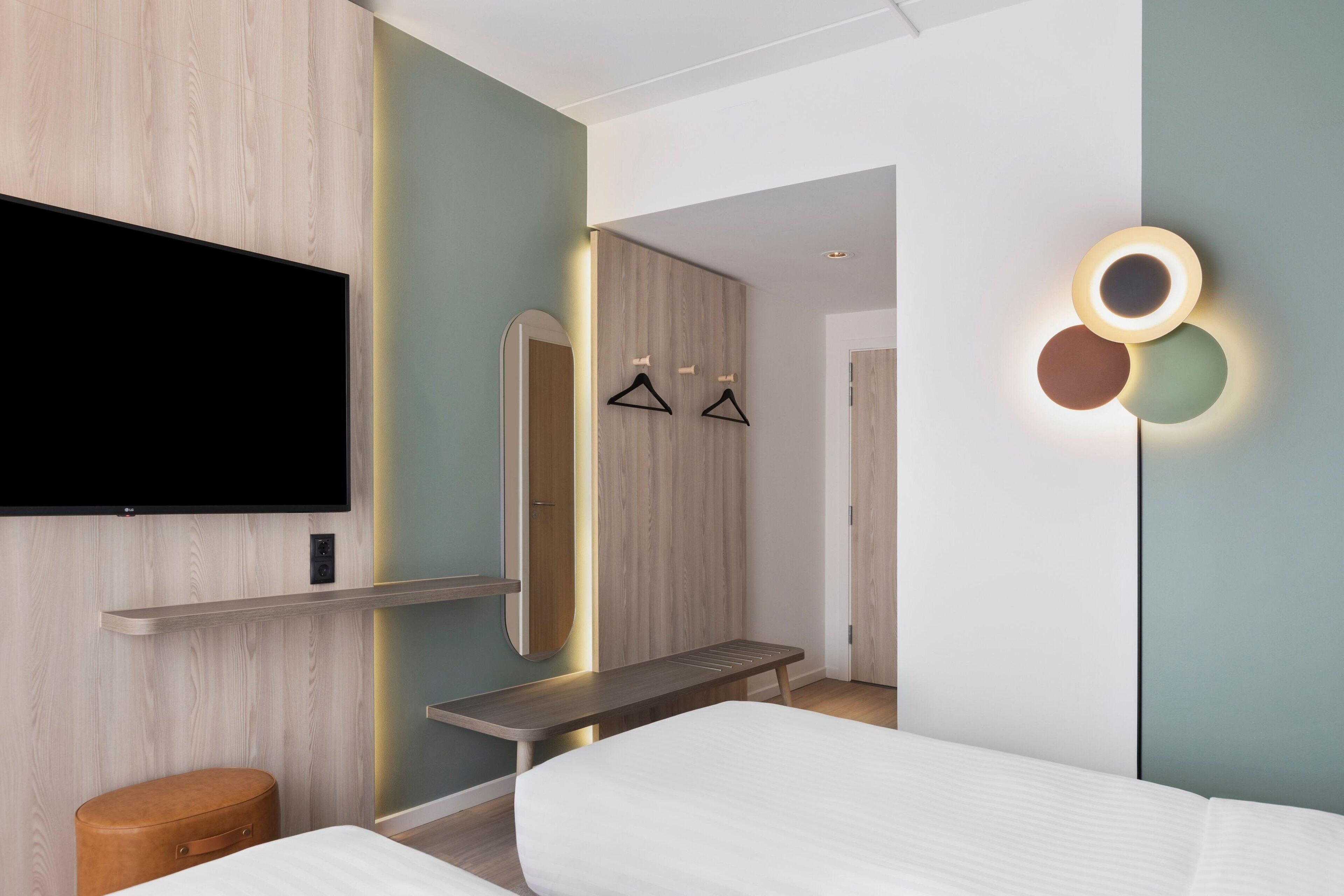 A modern hotel room with a flat-screen TV, a bench, a bed, and a wall-mounted mirror.