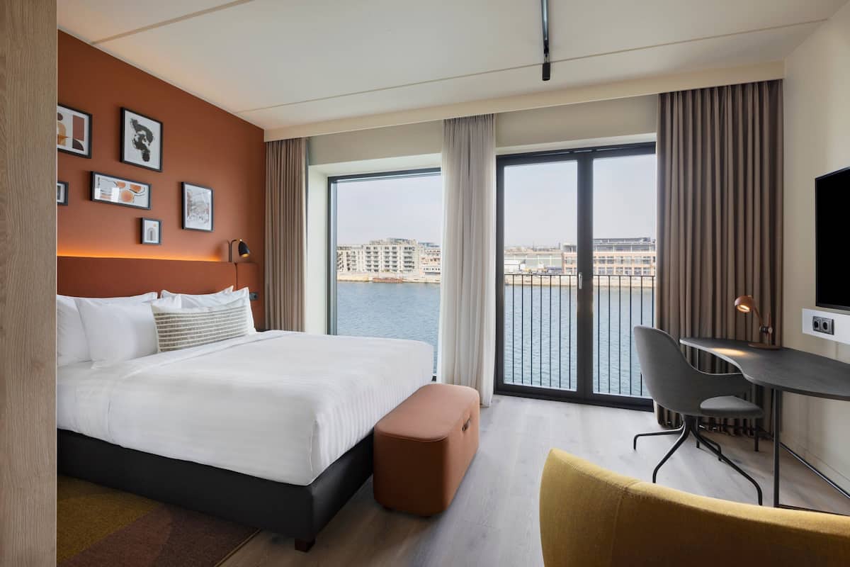 A modern hotel room with a large bed, a desk, a chair, and a view of the city through the window.