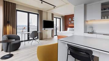 Une chambre d’hôtel moderne dotée d’une grande fenêtre, d’un téléviseur à écran plat, d’un lit, d’un petit coin cuisine et d’un espace salon avec une table et des chaises.