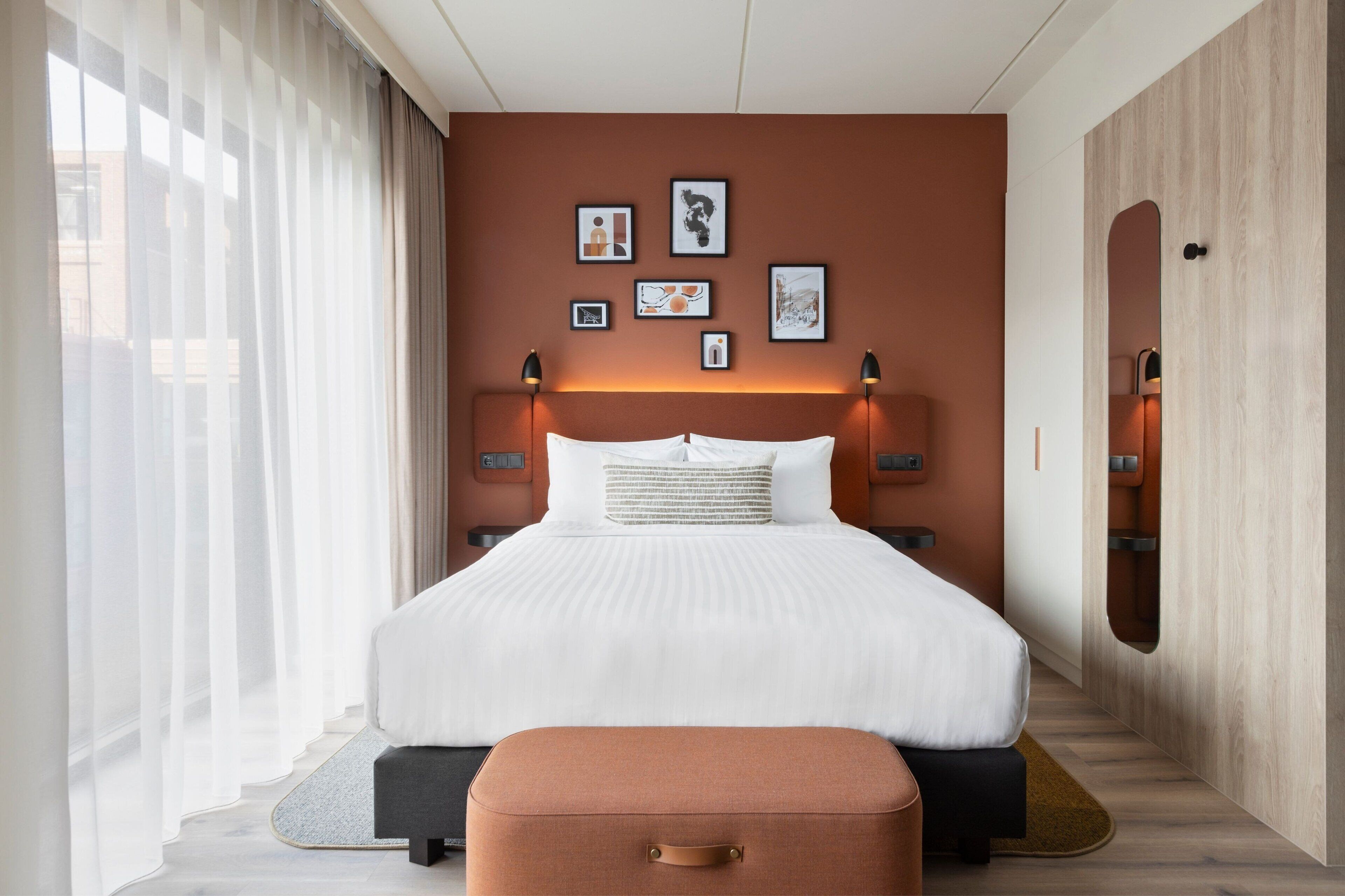 A modern hotel room with a large bed, a bedside table, a wall-mounted mirror, and a window with sheer curtains.