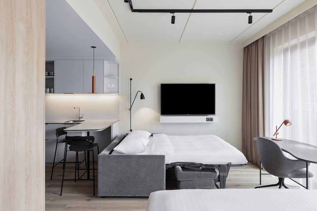 A modern hotel room with a bed, a desk with a chair, a TV mounted on the wall, and a window with curtains.