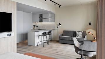 A modern hotel room with a flat-screen TV, a dining area with a table and chairs, a sofa, and a kitchenette.