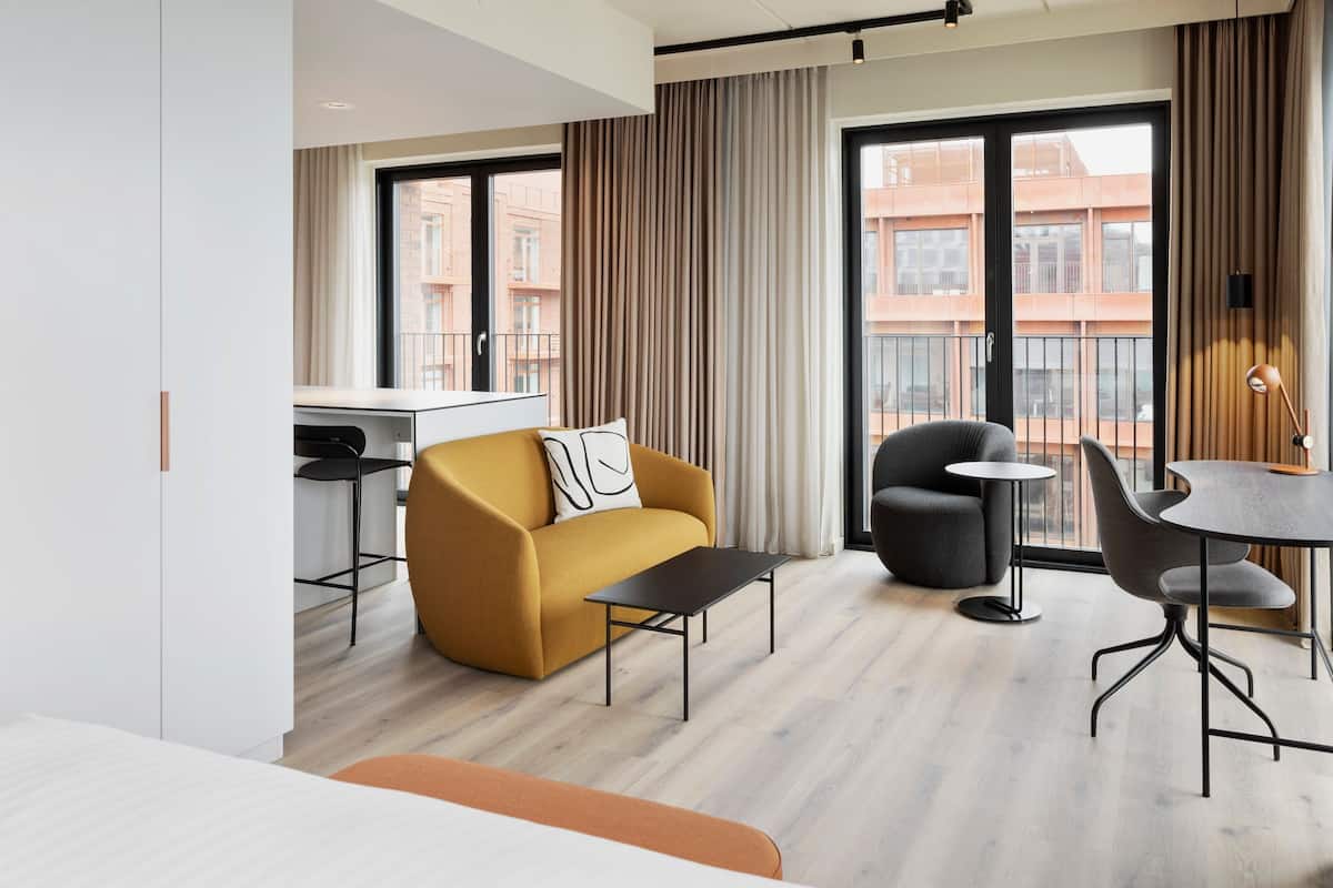 A modern hotel room with a sofa, a small table, a dining area, and a view of the cityscape through large windows.