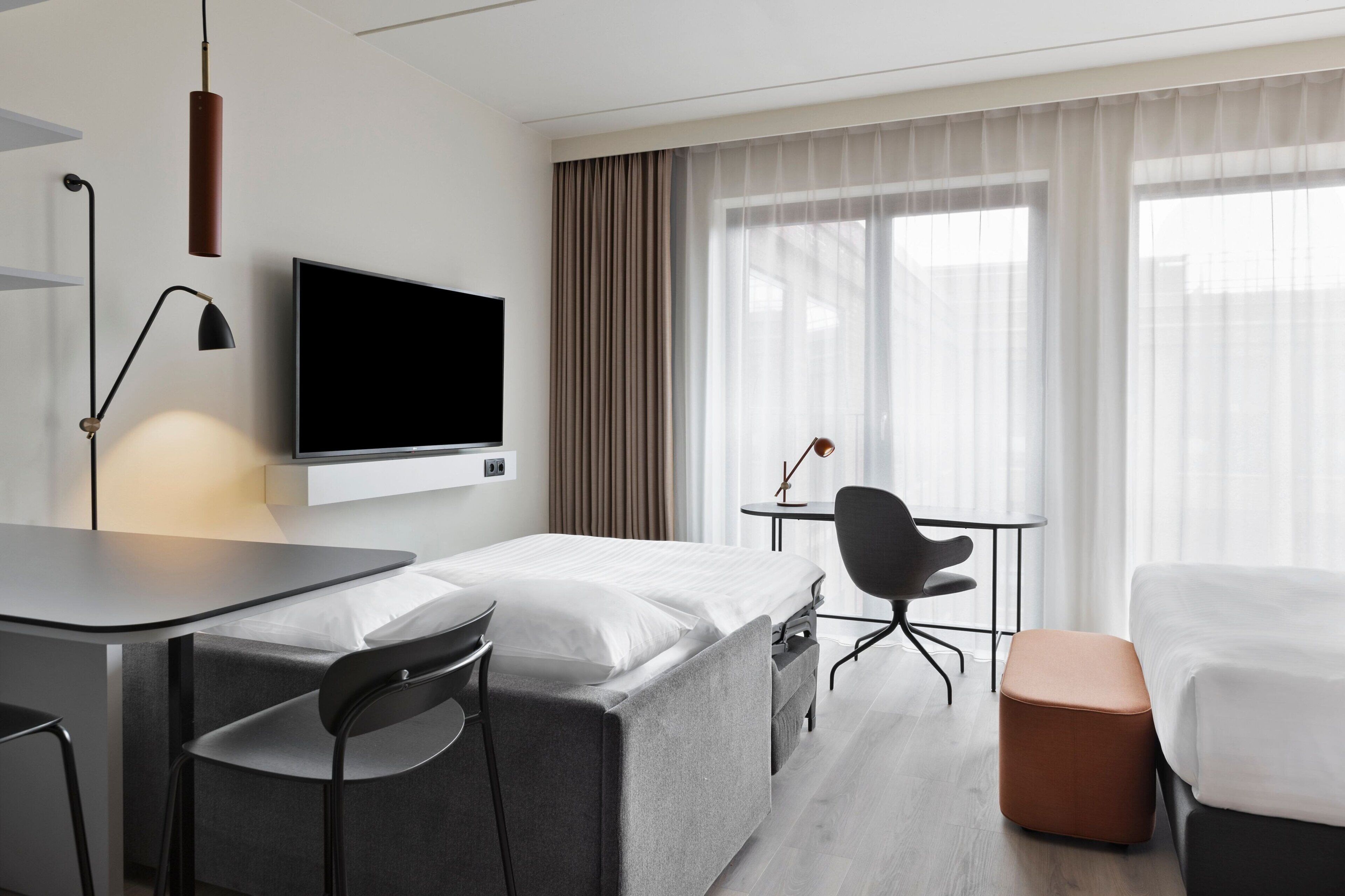 A hotel room with a bed, a desk, a chair, a TV, and a window with curtains.